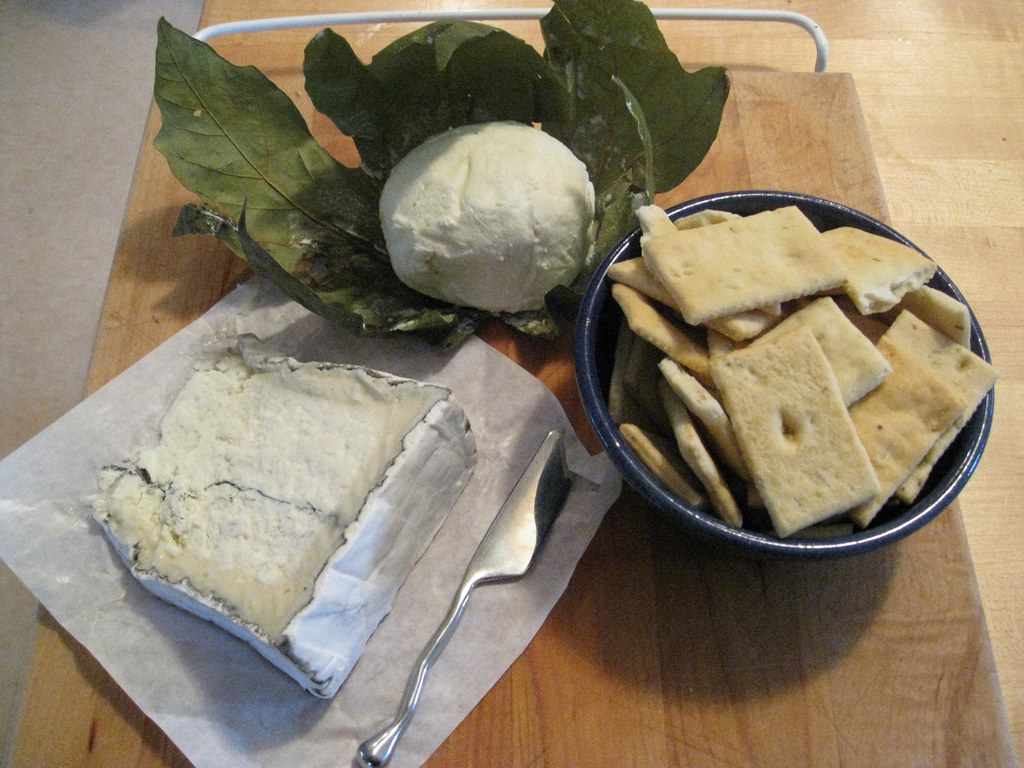 Humboldt Fog cheese board showing a wedge of soft cheese with a grey ash layer, a ball of soft cheese and a bowl of small square crackers
