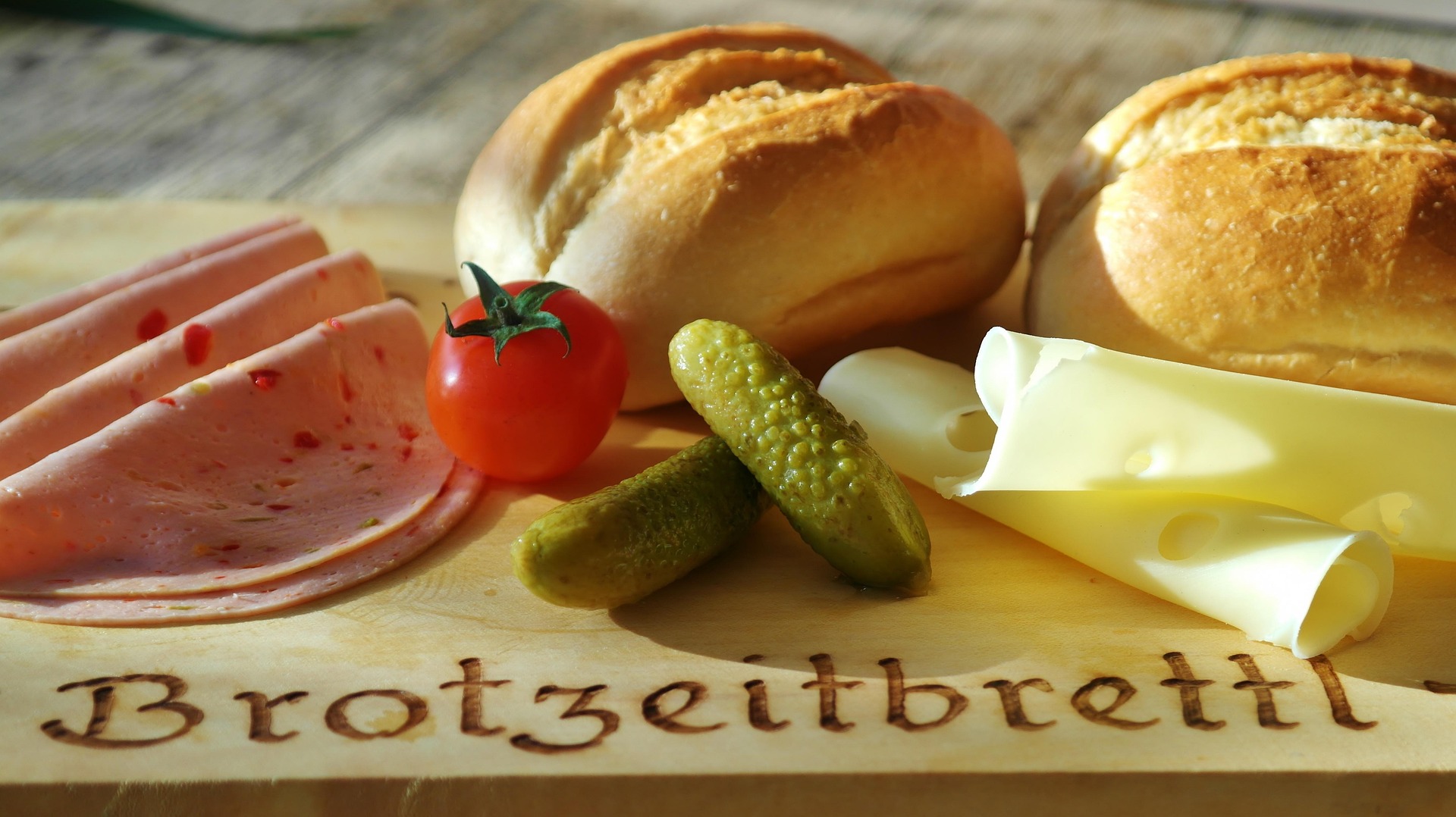 A cheeseboard is nothing without something pickled -- A cheese board containing crusty bread, some pale cheese slices, a tomato and several pickled cucumbers. The board is branded with Brotzeitbrettl
